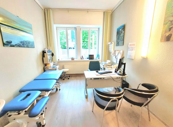 Heller Behandlungsraum mit blauer Behandlungsliege, Schreibtisch mit Computer und schwarzen Stühlen vor großem Fenster