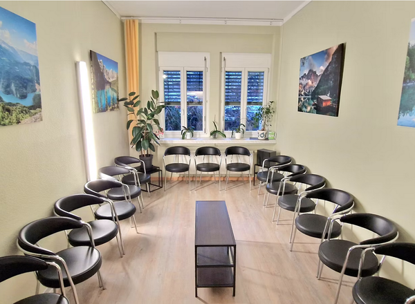 Wartezimmer mit schwarzen Stühlen im Kreis angeordnet, großformatige Naturbilder von Bergen und Seen an den Wänden