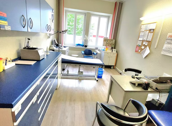 Moderner Behandlungsraum mit blauen Schränken, Behandlungsliege und hellem Fenster