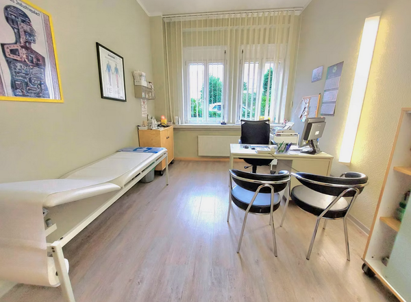 Heller Behandlungsraum mit weißer Untersuchungsliege, Schreibtisch mit Computer und zwei schwarzen Besucherstühlen vor großen Fenstern