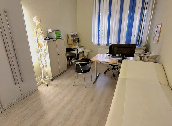 Moderner Behandlungsraum mit weißer Untersuchungsliege, Schreibtisch mit Computer und Skelettmodell an der Wand